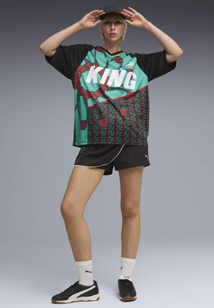 Noor naine, kes kannab mustriga "KING" spordisärki, musti lühikesi pükse, valgeid sokke, musti tossusid ja musta nokamütsi, poseerimas käed otsa ees.