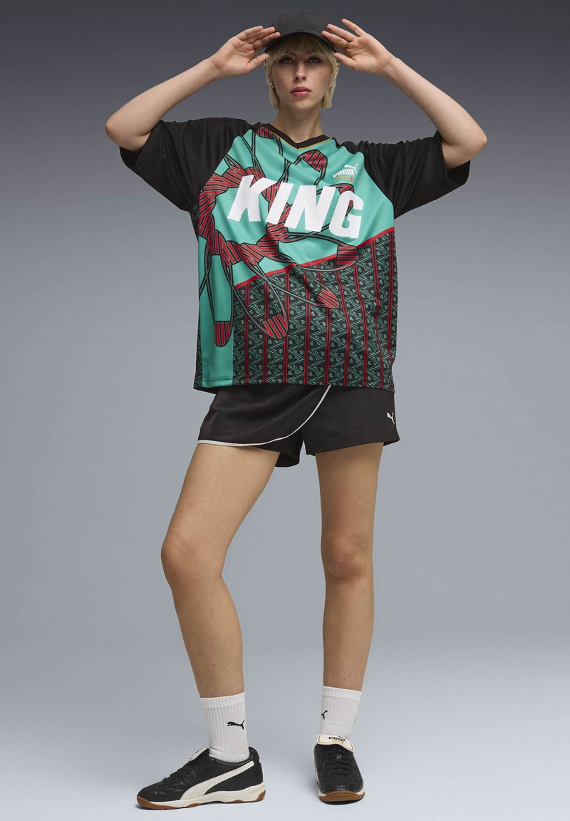 Νεαρή γυναίκα με αθλητική φανέλα με σχέδιο και τη λέξη "KING", μαύρο σορτς, άσπρες κάλτσες, μαύρα αθλητικά παπούτσια και μαύρο καπέλο, ποζάροντας με τα χέρια κοντά στο μέτωπο.