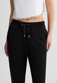 Pantaloni jogger neri con cintura regolabile a cordoncino, dotati di estremità dorate. Tessuto morbido che offre una vestibilità comoda e una texture liscia.