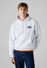 Felpa bianca in cotone con cappuccio e tasca frontale. Presenta un logo ricamato "Red Bull Racing" in blu sul petto. Jeans blu.