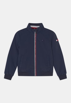 Marineblauwe jas met ritssluiting, opstaande kraag, twee voorzakken, geribde manchetten en zoom, en kleine rood-wit-blauwe vlaglogo's op borst en mouw.