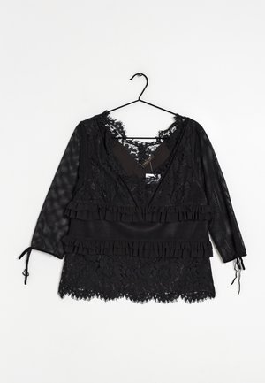 Blouse en dentelle noire avec manches trois-quarts en résille, détails à volants et fermetures à nouer aux poignets, suspendue sur un cintre noir sur fond blanc.