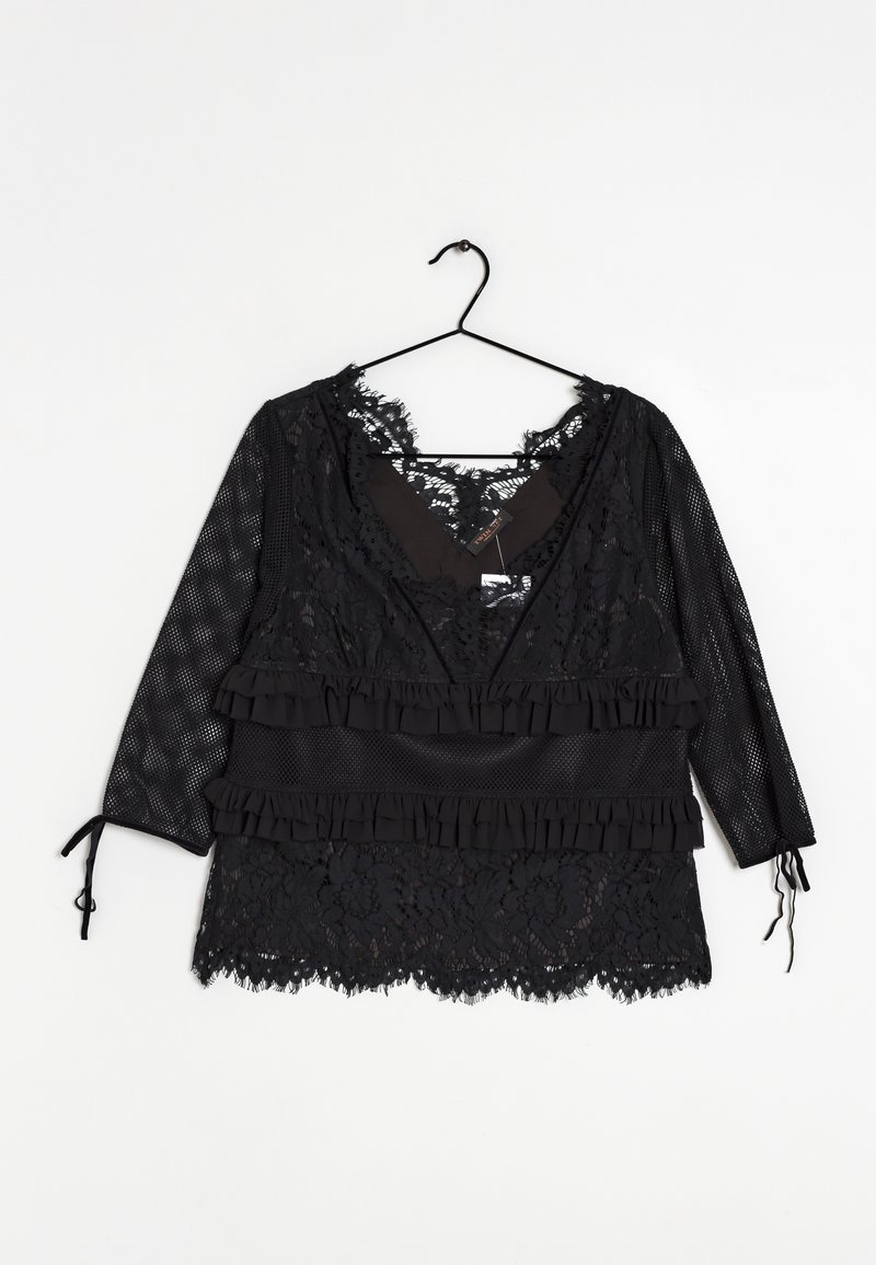 Blouse en dentelle noire avec manches trois-quarts en résille, détails à volants et fermetures à nouer aux poignets, suspendue sur un cintre noir sur fond blanc.
