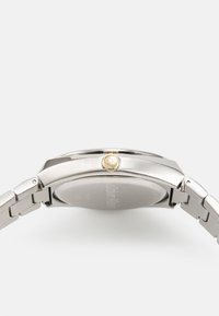 Reloj de pulsera de acero inoxidable con un acabado pulido, que presenta una corona en tono dorado y una caja redonda y suave. La correa es metálica y está articulada.
