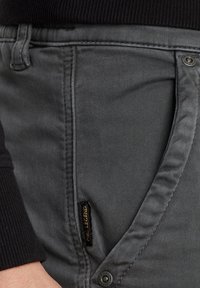 Graue Denim-Hosen mit einer glatten Textur, schmaler Passform, schrägen Seitentaschen und einem schwarzen Label mit goldener Schrift auf der Vorderseite.