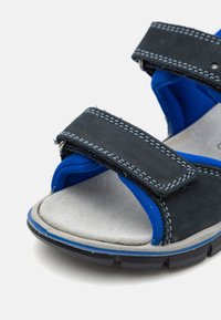 Schwarz-blaue Sandalen mit verstellbaren Klettverschlüssen, einer strukturierten grauen Innensohle und einer Gummisohle mit Rillen für besseren Halt.