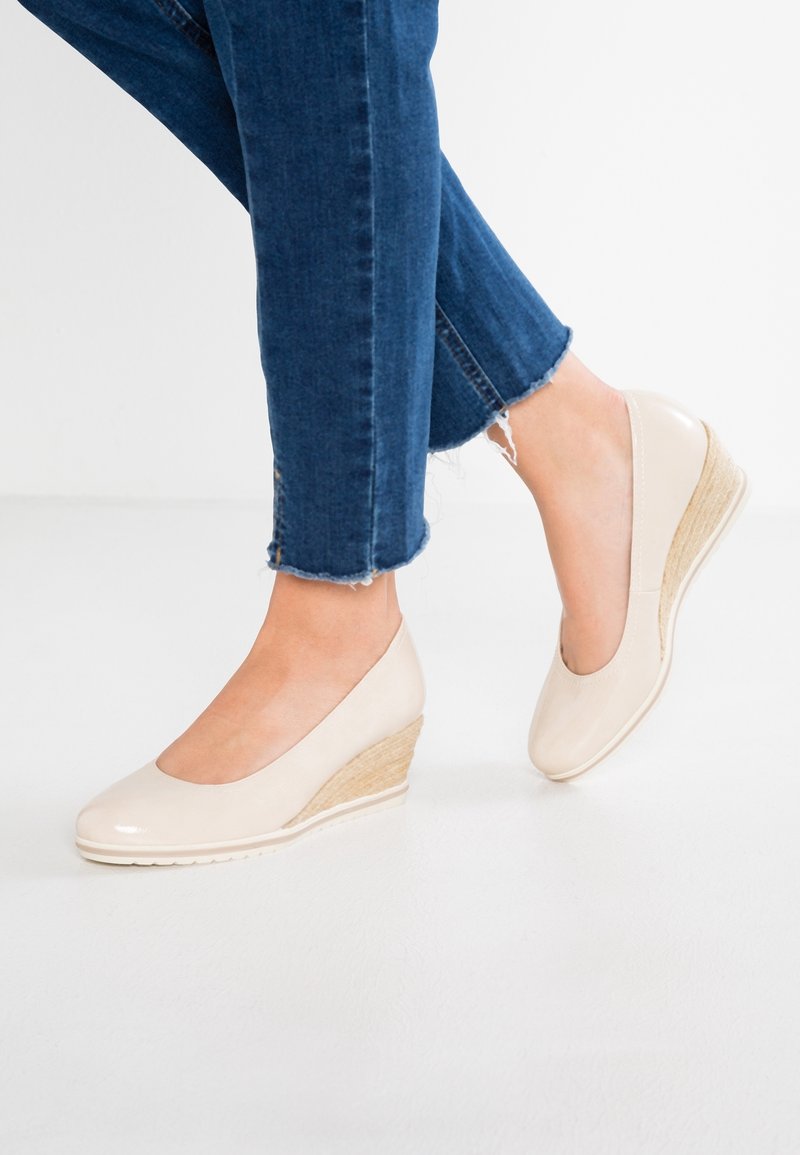 Pieds portant des talons compensés beige associés à un jean bleu denim raccourci avec des ourlets effilochés sur un sol blanc.