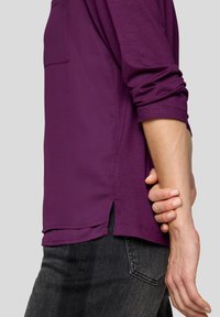 Lila Langarmshirt mit einer Tasche, mit aufgerollten Ärmeln und seitlichen Schlitzen. Getragen mit dunklen Jeans, die eine weiche, glatte Textur zeigen.