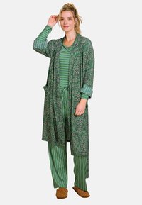 Groene met bloemenpatroon gewaad over gestreepte groene pyjama-set, met losse pasvorm, lange mouwen en zijzakken. Bruine slippers maken de look compleet.