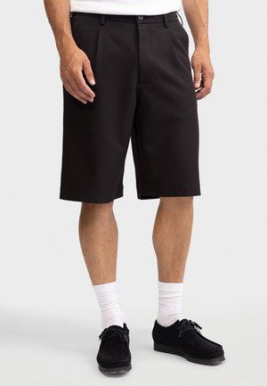 Homme portant un short noir arrivant aux genoux, des chaussettes blanches et des chaussures noires à lacets, se tenant devant un fond clair uni.