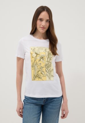 Young woman with long brown hair wearing white t-shirt with yellow botanical floral print and blue jeans, standing against plain background.