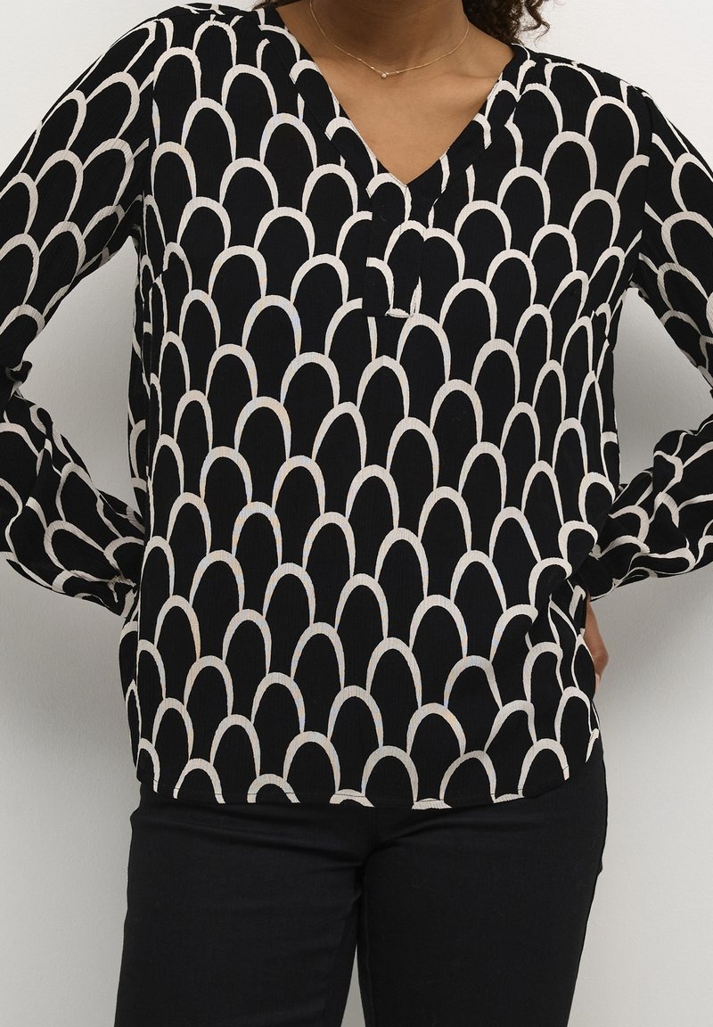 Femme portant un chemisier noir à manches longues avec un motif coquille blanc et un pantalon noir, les mains sur les hanches, sur un fond uni.