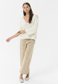 Pull tricot oversized beige clair avec un col en V et des manches larges, associé à un pantalon beige à jambes larges et des chaussures claires. Tissu simple et texturé.