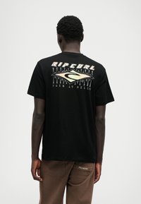 T-shirt de algodão preto com um grande gráfico nas costas com o texto "RIP CURL" e um logotipo. Usado com calças cargo castanhas.