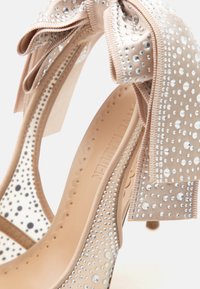 Μπεζ γυναικείο παπούτσι με ψηλό τακούνι διακοσμημένο με μικρές στρας και μεγάλο φιόγκο με στρας στη φτέρνα.