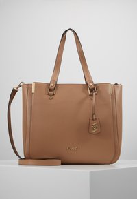Torebka typu tote w kolorze brązowym z ekoskóry, z podwójnymi uchwytami, odpinanym paskiem na ramię, złotymi akcentami oraz metalową zawieszką z logotypem. Gładka faktura.