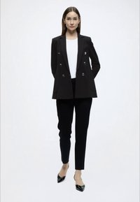Μαύρο σακάκι διπλής σειράς με ασημένια κουμπιά, σε συνδυασμό με εφαρμοστά μαύρα παντελόνια και μυτερά μαύρα τακούνια, πάνω από λευκή μπλούζα.