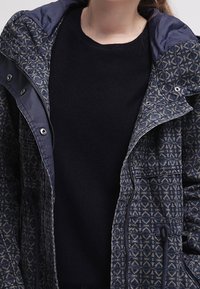 Marinblå mönstrad jacka i texturerat tyg, med huva och silverfärgade detaljer. Bärs över en enfärgad svart tröja. Har dragsko och fickor.