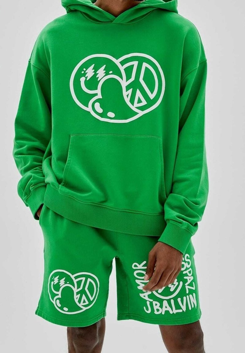 Hombre con sudadera verde brillante y pantalones cortos con diseños gráficos blancos que incluyen una cara sonriente, un corazón, un símbolo de paz y texto en los pantalones cortos.