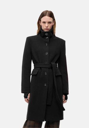 Persona che indossa un cappotto nero abbottonato con collo alto e cintura in vita, in piedi con le mani rilassate ai lati su uno sfondo semplice.