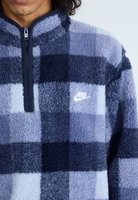 Pullover in fleece con zip a metà, caratterizzato da un motivo a quadri in diverse tonalità di blu e navy. Logo bianco sul petto, tessuto testurizzato.