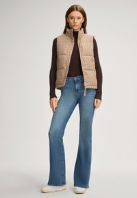 Beige gepolsterte Weste mit hohem Kragen und Druckknopfverschluss, kombiniert mit einem eng sitzenden dunkelbraunen Rollkragenpullover und weit geschnittenen Jeans.