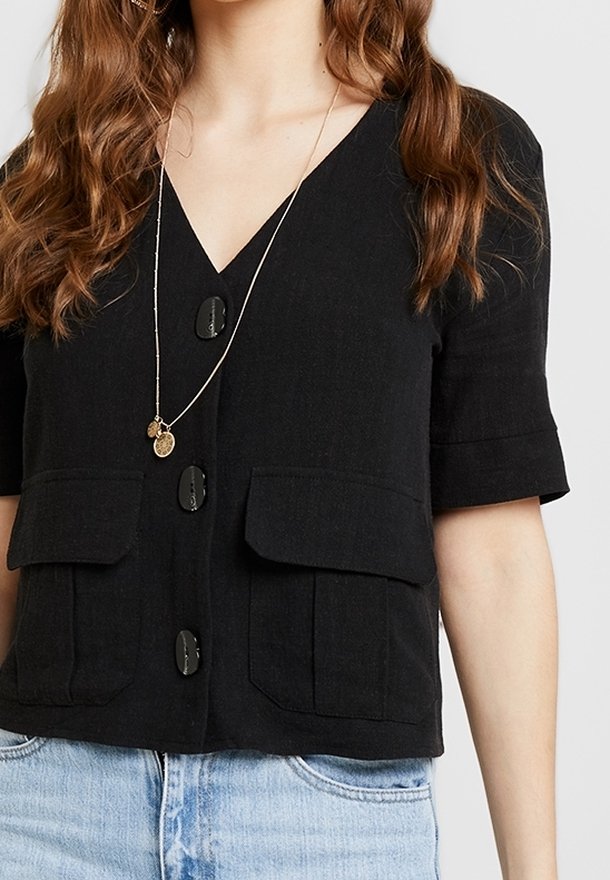Femme portant un cardigan noir à manches courtes boutonné avec des poches avant et un jean bleu clair, accessoirisée d'un collier pendentif en or.