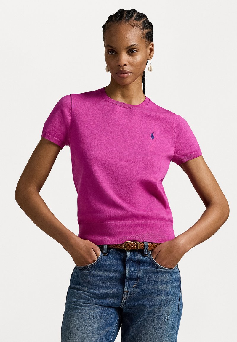 T-shirt en tricot rose ajusté à manches courtes avec un col rond et un petit logo bleu sur la poitrine. Assorti à un jean bleu taille mi-haute et une ceinture en cuir tressé marron.