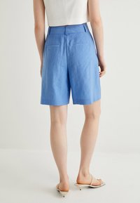 Blå linneshorts med avspänt snitt, utrustade med bakfickor och en jämn textur. Stylat med en vit kort topp och ljusa skor.