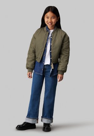 Ragazza sorridente con lunghi capelli scuri che indossa una giacca bomber verde, una camicia in denim blu e jeans, una maglietta bianca e scarpe nere, che si trova su uno sfondo grigio.