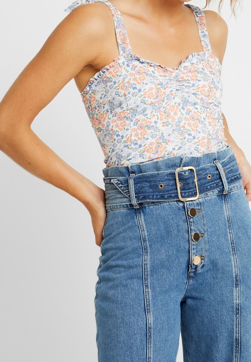 Femme portant un haut sans manches à fleurs rentré dans un jean bleu taille haute avec une large ceinture en denim et une boucle métallique, mains posées sur les hanches.