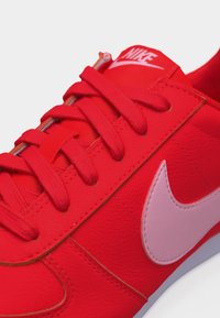Κόκκινο αθλητικό παπούτσι Nike με ροζ λογότυπο swoosh, κόκκινα κορδόνια και σήμανση Nike στη γλώσσα, σε κοντινό πλάνο.