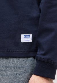 Μανίκι και τελείωμα μπλε ναυτικού φούτερ με λευκή ορθογώνια ετικέτα λογότυπο Hugo, σε συνδυασμό με μπλε-γκρι τζιν παντελόνι στο χέρι ενός ατόμου.