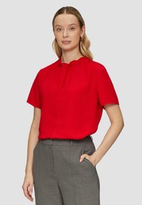 Rote kurzärmelige Bluse mit gerafftem Hals und subtilen Faltendetails, kombiniert mit grauen maßgeschneiderten Hosen. Glatte Stofftextur.