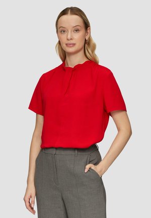 Rote kurzärmelige Bluse mit gerafftem Hals und subtilen Faltendetails, kombiniert mit grauen maßgeschneiderten Hosen. Glatte Stofftextur.