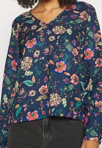 Blouse florale navy avec un col en V, ornée de motifs colorés de fleurs et de feuilles, manches longues et coupe décontractée. Texture de tissu lisse.