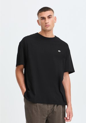 Giovane con capelli corti che indossa una t-shirt nera con un piccolo logo sul petto e pantaloni di velluto a coste marroni su sfondo bianco.