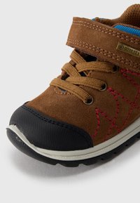 Chaussure pour enfant en daim marron avec embout en caoutchouc noir, couture rose, doublure bleue et bande velcro. Caractérisée par une semelle texturée pour une meilleure adhérence.