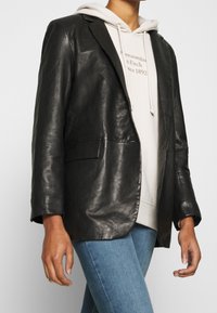 Schwarzer Lederblazer mit glatter Textur, Ein-Knopf-Verschluss und zwei vorderen Taschen, getragen über einem hellfarbigen Hoodie und Jeans.