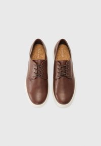 Chaussures en cuir marron avec une surface texturée, dotées de laçages et d'une semelle en caoutchouc blanc. Semelle intérieure en liège visible à l'intérieur.