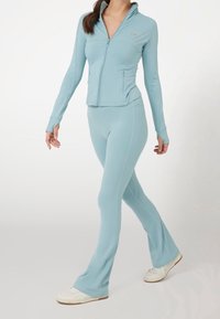 Giacca sportiva azzurra chiara con zip e maniche lunghe, abbinata a pantaloni a palazzo aderenti dello stesso colore. Il materiale sembra liscio ed elastico. Sneakers bianche.