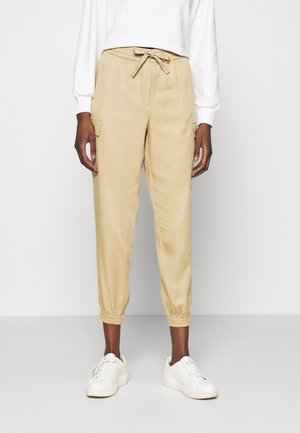 Osoba ubrana w beżowe spodnie dresowe cargo z elastycznymi mankietami i ściągaczem w talii, w połączeniu z białymi trampkami i białą bluzką z długim rękawem.