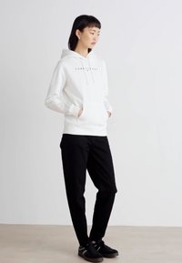 Sudadera blanca con cordones y bolsillos frontales, que presenta un logo simple. Combinada con pantalones negros y zapatos negros. Textura de tela suave.