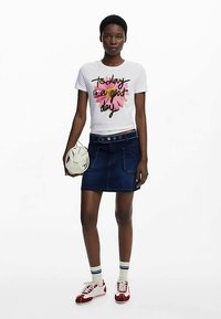 Wit T-shirt met bloemmotief en zwarte letters, gecombineerd met een donkere denimrok. Accessoires zijn onder andere een witte tas en rode sneakers.