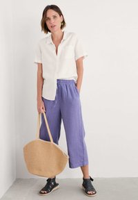 Camicia bianca a maniche corte con bottoni, pantaloni cropped in lino viola, sandali neri con cinturini e una grande borsa a tracolla in paglia beige.