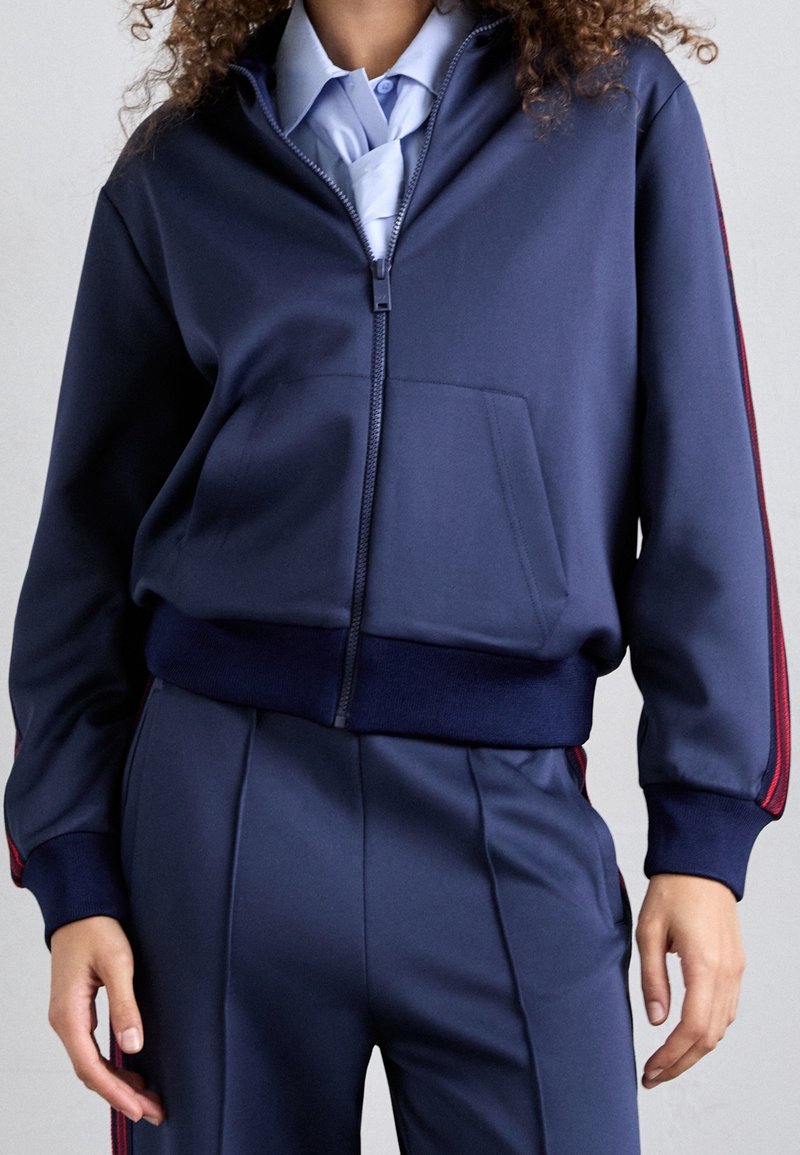 Tmavě modrá bunda na zip s volnějším střihem, s přední kapsou, žebrovanými manžetami a červenými akcentními pruhy podél rukávů.