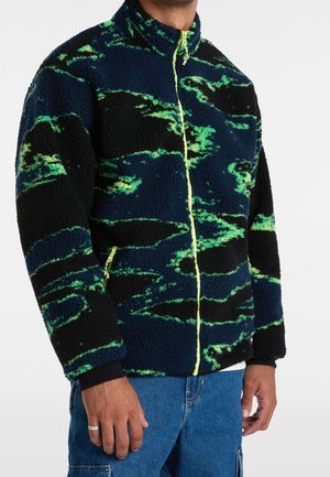 Person trägt eine Fleecejacke in Marineblau, Schwarz und Grün mit einem neongelben Reißverschluss, kombiniert mit blauen Jeanshosen.