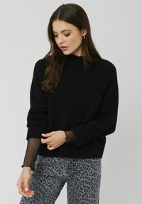 Pull en tricot noir avec un col montant et des poignets côtelés, superposé à une longue manche en mesh noir transparent. Assorti à un jean gris à motif animal.