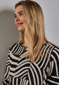 Musta ja kerma raidallinen paita, jossa on aaltoileva geometrinen kuvio, pyöreä pääntie ja pitkät hihat, yhdistettynä löysään hiustyyliin harmaata seinää vasten.