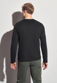 Mustat pitkät hihat -paita, jossa on pyöreä kaula-aukko, nähtynä takaa. Kangas näyttää sileältä, ja se on yhdistetty oliivinvihreisiin housuihin.